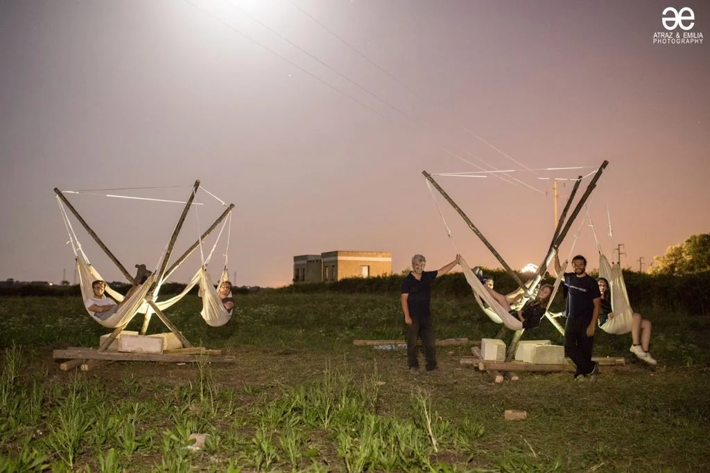 The two finished frames lit at night in the field, people resting in the hammocks, the glow of the sky behind them